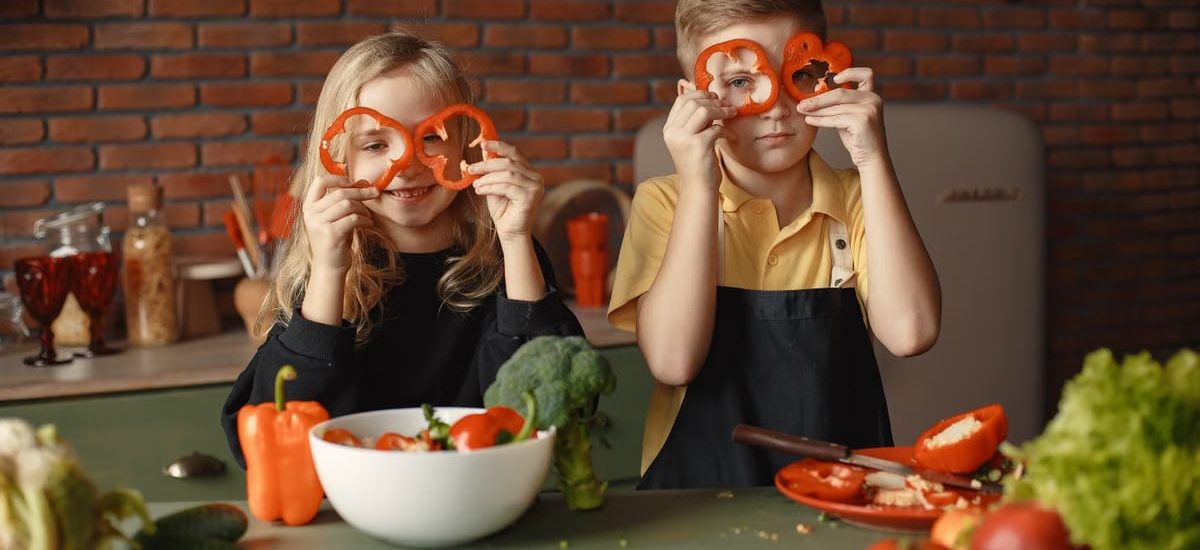 Funny boy and girl wearing apron looking through slices of pepper while preparing healthy vegetarian salad together at table with ingredients and bowls