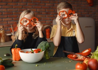 Funny boy and girl wearing apron looking through slices of pepper while preparing healthy vegetarian salad together at table with ingredients and bowls