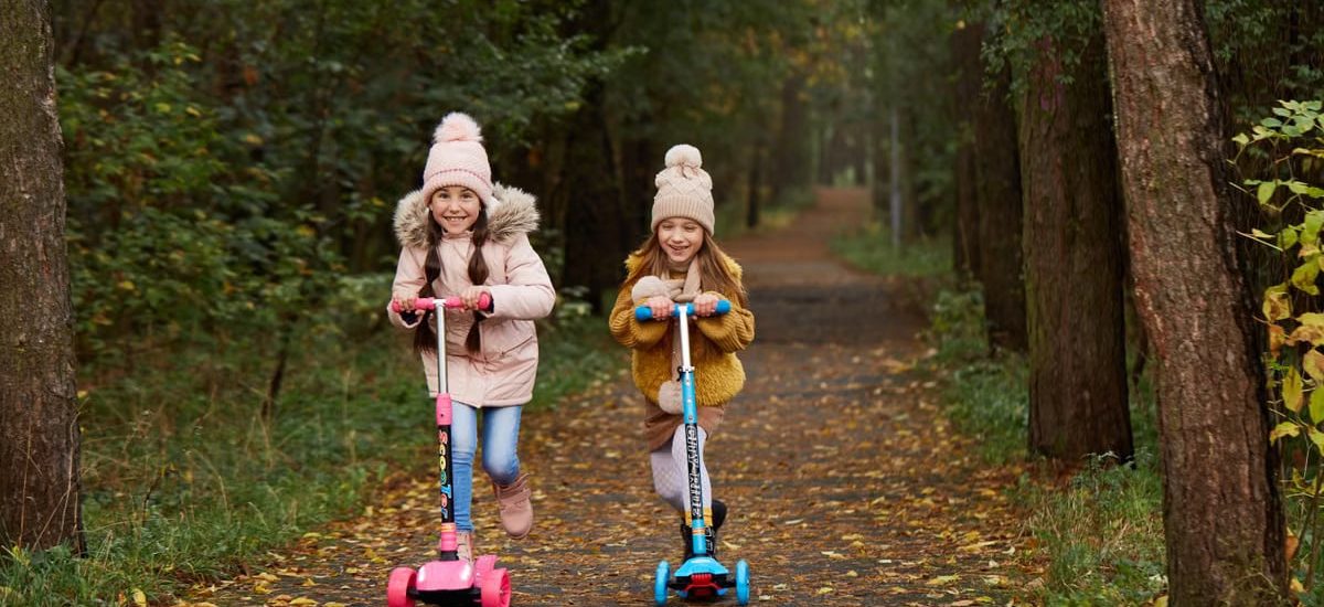 Two girls wearing beanies and winter jackets ride scooters on a leaf-strewn path in an autumn park.
