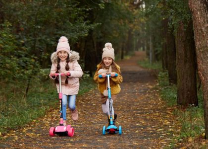 Two girls wearing beanies and winter jackets ride scooters on a leaf-strewn path in an autumn park.