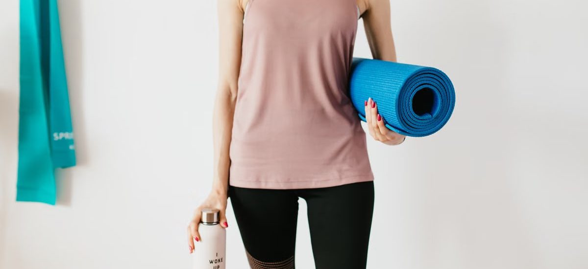 Faceless slim female athlete in sportswear standing with blue fitness mat and water bottle while preparing for indoors workout